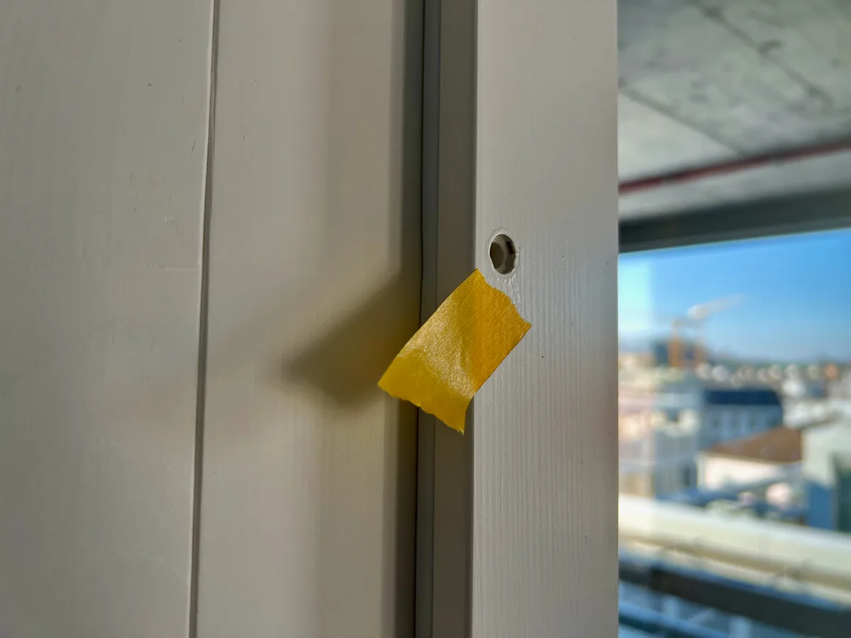 Ein gelber Klebestreifen klebt neben einem Loch in einem Holzrahmen, in dem die Schraube fehlt.